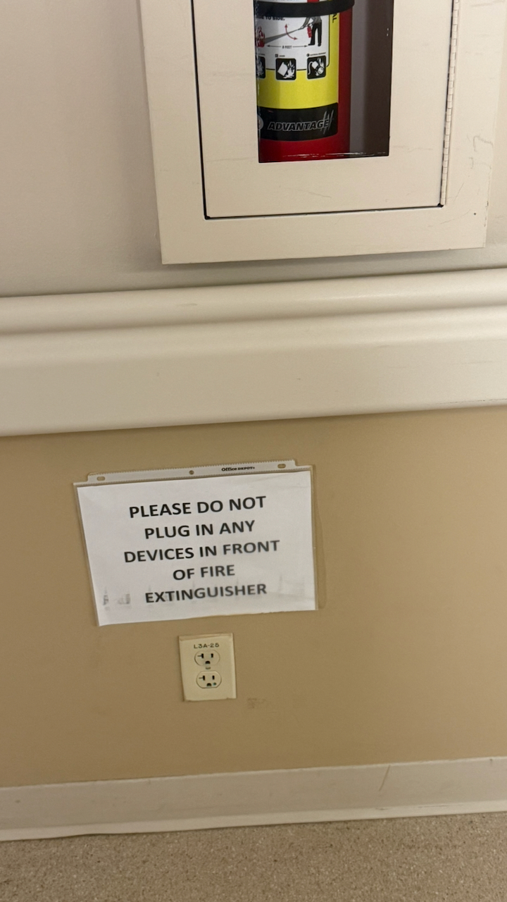 A hospital wall outlet beneath a paper sign reading, “Please do not plug in any devices in front of fire extinguisher.”